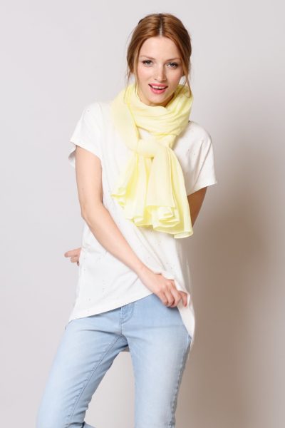Une mannequin aux cheveux longs châtain clair attachés en arrière porte un t-shirt blanc ample, un jean bleu clair et un foulard jaune clair. Elle sourit et bouge légèrement, une main sur la hanche et l'autre tenant une partie de sa chemise. L'arrière-plan évoque le chic parisien avec son gris clair uni, une capture parfaite par un photographe de mode.