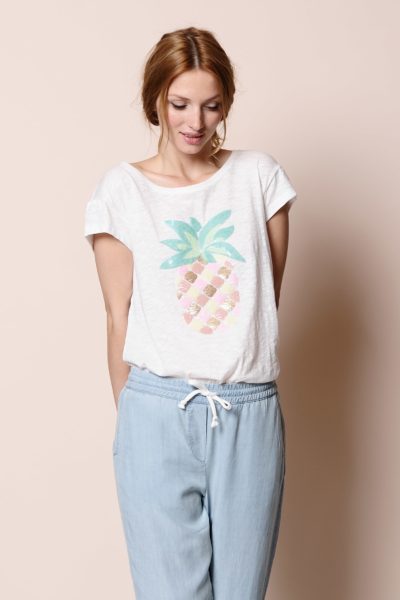 Une mannequin aux cheveux châtain clair porte un t-shirt blanc avec un grand motif d'ananas coloré sur le devant. Elle l'associe à un pantalon bleu clair à cordon. Debout sur un fond beige uni, elle regarde vers le bas avec un léger sourire, dégageant une simplicité de mode parisienne que tout photographe admirerait.