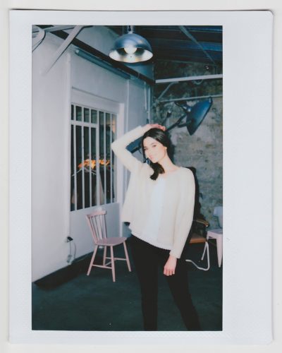 Une mannequin aux cheveux longs et noirs, vêtue d'un pull clair et d'un pantalon noir, pose en tenant ses cheveux relevés d'une main. Elle se tient debout dans une pièce faiblement éclairée par un éclairage industriel, un mur blanc et une chaise rose en arrière-plan. La photo, du photographe parisien Thomas Duclert, a une bordure de style polaroïd.