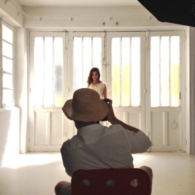 Le photographe Thomas Duclert, coiffé d'un chapeau de paille, prend des photos d'une mannequin debout devant une grande porte en verre dépoli dans une pièce blanche bien éclairée. La mannequin fait face à l'objectif et la lumière du soleil illumine l'espace.