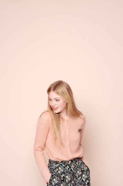 Une mannequin aux longs cheveux blonds se tient debout sur un fond beige. Elle porte un cardigan rose clair avec une broche et un pantalon taille haute à motifs floraux. Dans le style parisien, elle regarde vers la gauche, les mains dans les poches, souriant légèrement.
