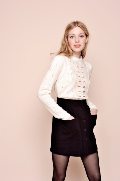 Une mannequin aux longs cheveux blonds, vêtue d'un pull blanc à manches longues avec des motifs découpés et un décolleté haut, associé à une jupe noire et des collants noirs, incarne la mode parisienne. Elle a les mains dans les poches de sa jupe et se tient debout sur un fond beige uni.