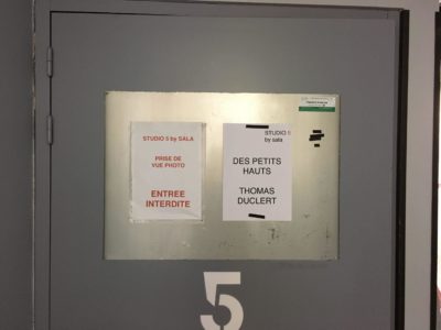 Une porte grise marquée "5" avec deux pancartes en papier collées dessus. La pancarte de gauche indique "PRISE DE VUE PHOTO, ENTREE INTERDITE" en français, et la pancarte de droite indique "STUDIO 5 by sala, DES PETITS HAUTS, Thomas Duclert". Un panneau d'urgence est placé au-dessus de la pancarte de droite. Photographe Paris mentionné ci-dessous.
