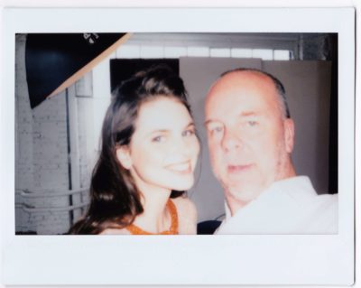 Une mannequin aux longs cheveux noirs et au grand sourire se tient à côté de Thomas Duclert. Ils prennent un selfie dans un studio de photographie avec des murs en briques et de grandes fenêtres en arrière-plan. L'image a une bordure blanche de style Polaroïd, reflétant parfaitement leur mode.