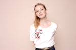 Une mannequin aux longs cheveux blonds, vêtue d'un t-shirt blanc à motif floral brodé, pose avec assurance sur un fond beige. Les mains derrière le dos et un jean en denim foncé associé à une ceinture beige, elle incarne la mode moderne, un moment capturé avec brio par le photographe Thomas Duclert.