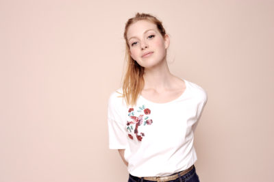 Une mannequin aux longs cheveux blonds, vêtue d'un t-shirt blanc à motif floral brodé, pose avec assurance sur un fond beige. Les mains derrière le dos et un jean en denim foncé associé à une ceinture beige, elle incarne la mode moderne, un moment capturé avec brio par le photographe Thomas Duclert.