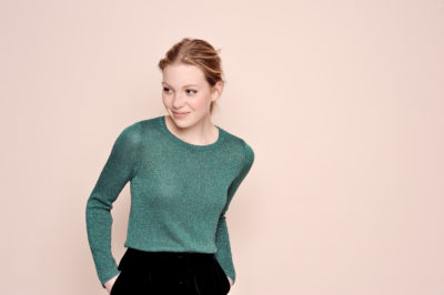 Une personne aux cheveux clairs se tient debout, les mains dans les poches, le regard légèrement sur le côté. Elle porte un pull vert à manches longues et un pantalon noir. L'arrière-plan est d'une couleur unie, beige clair, une scène qui rappelle l'esthétique de la mode parisienne de Thomas Duclert.