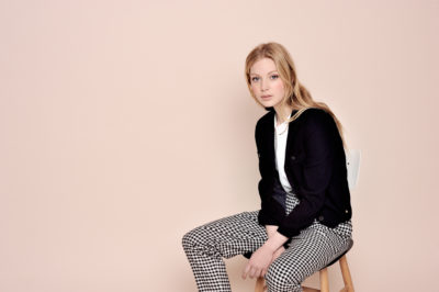 Une personne aux cheveux longs et blonds est assise sur un tabouret en bois sur fond beige. Elle porte une veste noire, une chemise blanche et un pantalon à carreaux noirs et blancs, le regard légèrement sur le côté avec une expression calme, comme l'a capturé le photographe Thomas Duclert.