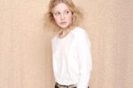 Une personne aux cheveux blonds ébouriffés, portant une chemise blanche à manches longues et une ceinture beige clair, se tient légèrement sur le côté, regardant au loin, comme si elle était capturée dans un cadre artistique du célèbre photographe Thomas Duclert sur un fond texturé neutre.