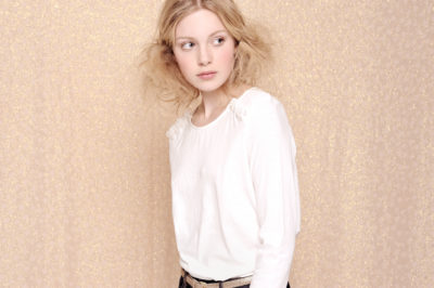 Une personne aux cheveux blonds ébouriffés, portant une chemise blanche à manches longues et une ceinture beige clair, se tient légèrement sur le côté, regardant au loin, comme si elle était capturée dans un cadre artistique du célèbre photographe Thomas Duclert sur un fond texturé neutre.