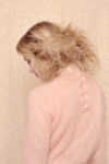 Une personne aux cheveux blonds volumineux se tient debout, le regard tourné vers le bas. Elle porte un pull rose pâle à manches longues avec des boutons dans le dos. Le décor respire la mode parisienne sur un fond beige clair moucheté.