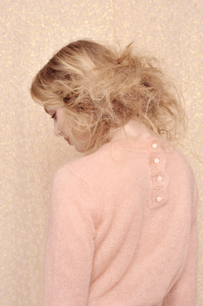 Une personne aux cheveux blonds volumineux se tient debout, le regard tourné vers le bas. Elle porte un pull rose pâle à manches longues avec des boutons dans le dos. Le décor respire la mode parisienne sur un fond beige clair moucheté.