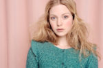 Une mannequin aux longs cheveux blonds, ébouriffés et légèrement ondulés, se tient devant un rideau rose. Elle porte un haut en tricot bleu canard avec des boutons. Son expression est neutre et son regard est doux et naturel – une capture classique du photographe de mode Thomas Duclert.