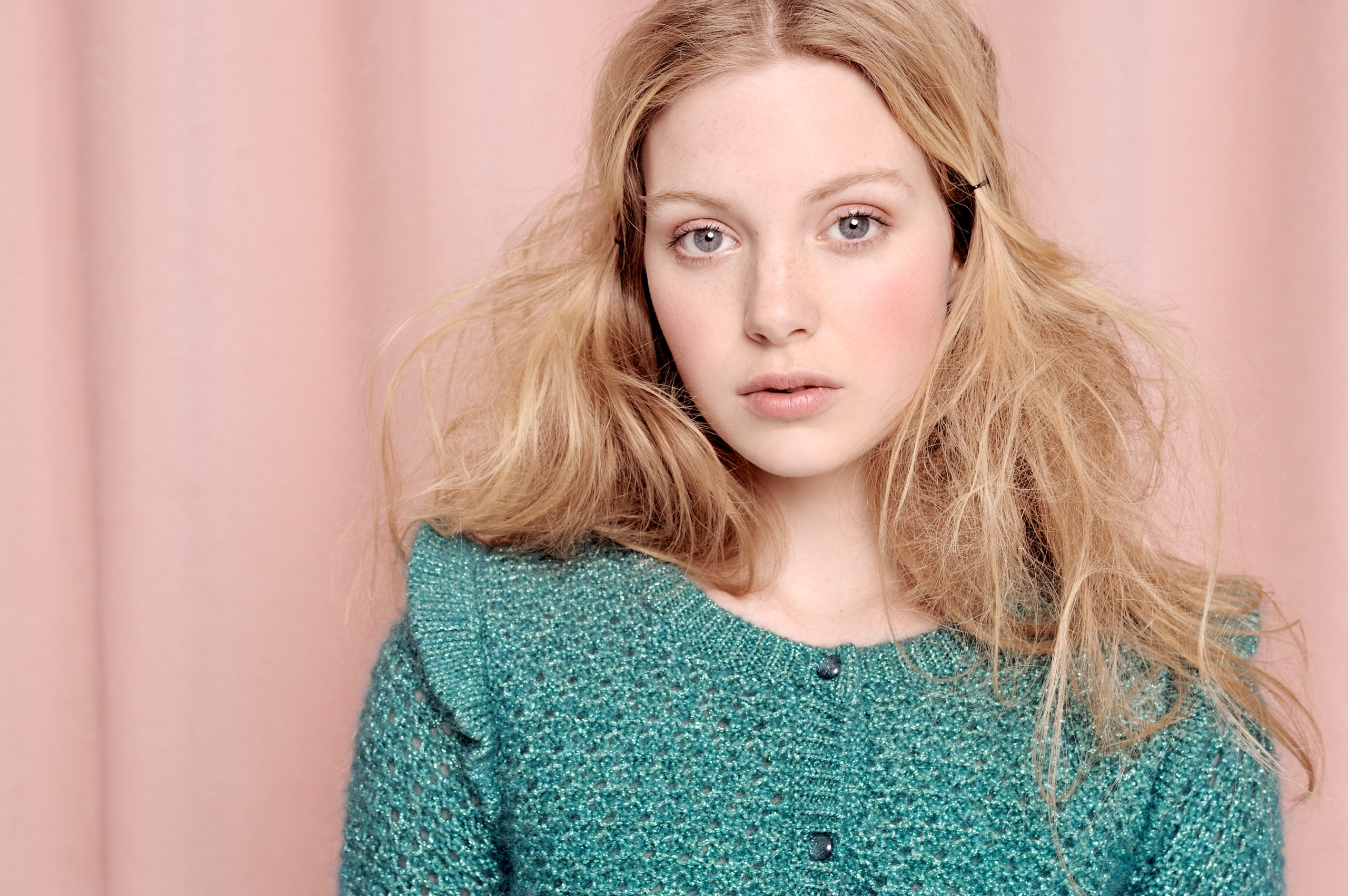 Une mannequin aux longs cheveux blonds, ébouriffés et légèrement ondulés, se tient devant un rideau rose. Elle porte un haut en tricot bleu canard avec des boutons. Son expression est neutre et son regard est doux et naturel – une capture classique du photographe de mode Thomas Duclert.