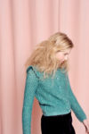 Une personne aux cheveux blonds longs et ondulés regarde vers le bas, debout devant un rideau rose. Photographiée par Thomas Duclert à Paris, elle porte un cardigan en tricot bleu canard avec des épaules à volants et un pantalon noir. La scène est doucement éclairée, créant une atmosphère douce.