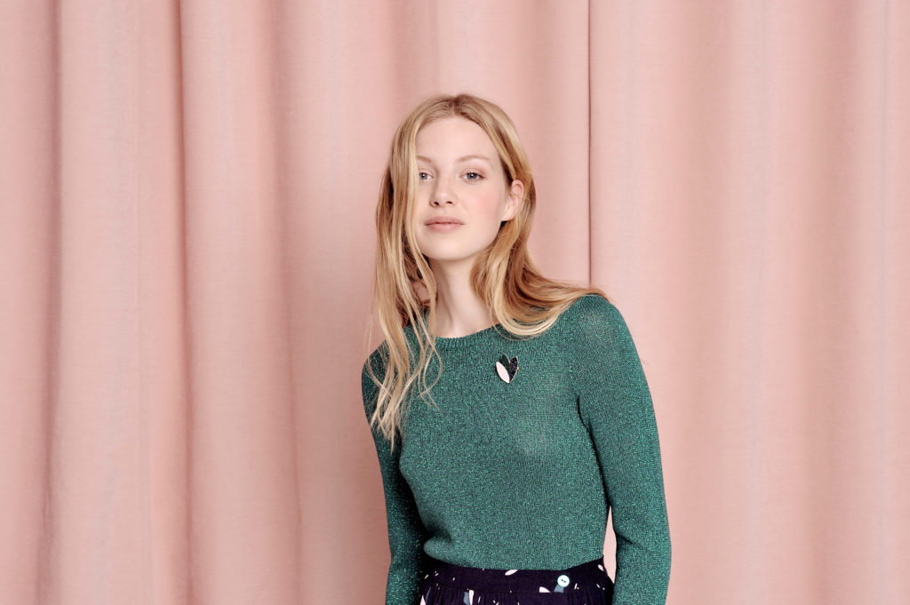 Une mannequin aux longs cheveux blonds se tient debout devant un rideau rose. Elle porte un haut vert à manches longues orné d'une broche en forme de cygne et une jupe à motifs mouchetés de blanc. Photographiée à Paris, elle dégage une expression détendue et confiante qui convient à tous les styles.
