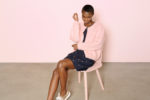 Une personne assise sur une chaise rose clair sur un fond rose clair. Elle porte un cardigan rose sur une robe sombre avec de petits ornements dorés et des baskets blanches. La personne, photographiée par le célèbre photographe parisien Thomas Duclert, a les cheveux courts et semble ajuster sa boucle d'oreille.