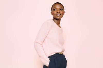 Une personne aux cheveux courts se tient debout sur un fond rose clair, souriant et regardant sur le côté tout en portant un pull rose clair et un pantalon foncé. Ses mains sont dans ses poches, capturant un moment chic sans effort, comme si elle était posée par le photographe de mode Thomas Duclert.