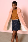 Une personne aux cheveux courts porte un manteau de fourrure couleur pêche sur un pull marron et un short bleu à taille haute avec un petit motif clair. Le fond est rose uni. La personne pose avec une jambe légèrement en avant et la tête légèrement inclinée vers le bas, mode capturée par le photographe Thomas Duclert.