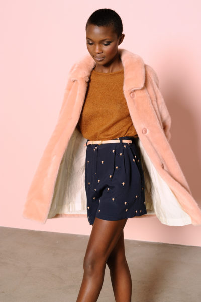Une personne aux cheveux courts porte un manteau de fourrure couleur pêche sur un pull marron et un short bleu à taille haute avec un petit motif clair. Le fond est rose uni. La personne pose avec une jambe légèrement en avant et la tête légèrement inclinée vers le bas, mode capturée par le photographe Thomas Duclert.