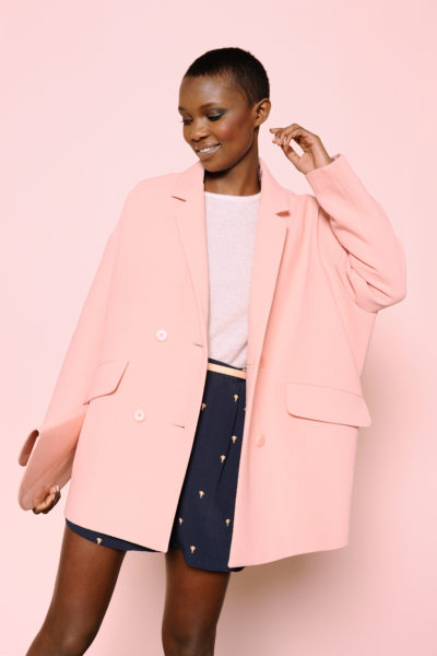 Une personne aux cheveux courts sourit tout en portant un blazer oversize rose clair, un haut rose clair et un short bleu marine à petits motifs dorés. Capture du photographe parisien Thomas Duclert, le fond est rose clair, complétant les couleurs de la tenue.