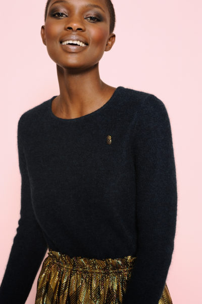 Une personne aux cheveux courts, vêtue d'un haut sombre à manches longues avec une petite broche, se tient debout sur un fond couleur pêche. Elle porte également une jupe à motifs dorés et noirs, évoquant la mode chic de Paris. La personne sourit et regarde légèrement ailleurs que devant la caméra.