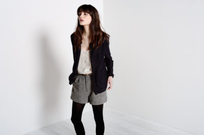 Une mannequin aux longs cheveux bruns se tient debout sur un fond blanc. Elle respire la mode parisienne avec un cardigan sombre, un haut blanc, un short taille haute gris et des collants noirs. Ses mains sont dans ses poches tandis qu'elle regarde légèrement sur le côté, évoquant un sentiment de minimalisme rappelant le style de Thomas Duclert.