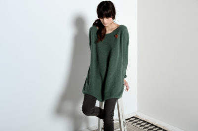 Une mannequin aux cheveux longs et noirs est assise sur un tabouret blanc contre un mur gris clair, incarnant la mode parisienne. Elle porte un pull vert oversize, un jean foncé et des bottes noires. Sa tête est légèrement inclinée vers le bas et sa main gauche repose sur le tabouret. Un tapis à motifs de Thomas Duclert est partiellement visible sur le sol.