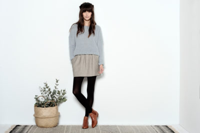 Une mannequin aux cheveux longs et bruns, vêtue d'un pull gris, d'une jupe à motifs, de collants noirs et de bottines marron, se tient debout devant un mur blanc uni. Elle pose avec une jambe croisée devant l'autre. Un panier rempli de verdure est posé sur le sol à côté d'elle. Photo prise par le photographe Thomas Duclert.