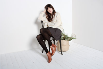 Une personne aux cheveux longs est assise sur une chaise noire sur fond blanc, rappelant une scène de la mode parisienne. Elle pose sa tête sur une main, porte un pull blanc, une jupe noire, des leggings noirs et des bottines marron. À côté d'elle, un panier tressé contenant des plantes vertes ajoute une touche d'élégance naturelle à la composition.