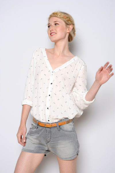 Une mannequin aux cheveux clairs se tient debout sur un fond uni, vêtue d'une chemise blanche à manches longues boutonnée avec des motifs à pois noirs, d'un short gris et d'une ceinture beige. Elle sourit et regarde sur le côté, incarnant sans effort la mode parisienne comme si elle était capturée par un photographe.