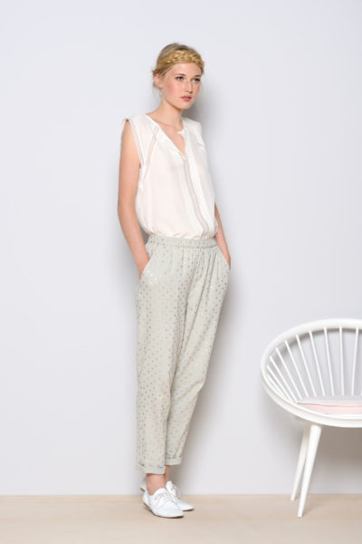 Une mannequin aux cheveux blonds se tient debout contre un mur gris clair. Elle porte un chemisier blanc sans manches, un pantalon gris clair à motifs discrets et des chaussures blanches. Elle a les mains dans les poches et regarde vers l'objectif. Une chaise blanche est placée à côté d'elle, capturant l'essence du style mode photographe de Thomas Duclert.
