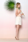 Une mannequin aux cheveux blonds se tient de profil sur un fond dégradé rose et blanc, tenant une grande feuille de palmier sur son épaule. Vêtue d'un haut sans manches rose clair, d'une jupe à pois blancs et de sandales beiges, elle respire le chic du style parisien à la Thomas Duclert.