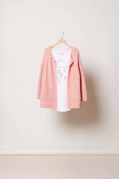 Un cardigan en tricot rose est suspendu à un cintre en bois contre un mur de couleur claire. Sous le cardigan, un t-shirt blanc avec un simple dessin au trait noir représentant un nuage et des gouttes de pluie est également suspendu. Photographiée par Thomas Duclert, cette image capture la simplicité parisienne sur un fond épuré.