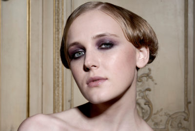 Une mannequin aux cheveux blonds courts et coiffés et aux yeux maquillés de noir regarde directement l'objectif avec une expression neutre. L'arrière-plan, rappelant l'élégance parisienne, présente un mur décoratif orné de couleur claire, une scène capturée avec brio par le photographe Thomas Duclert.