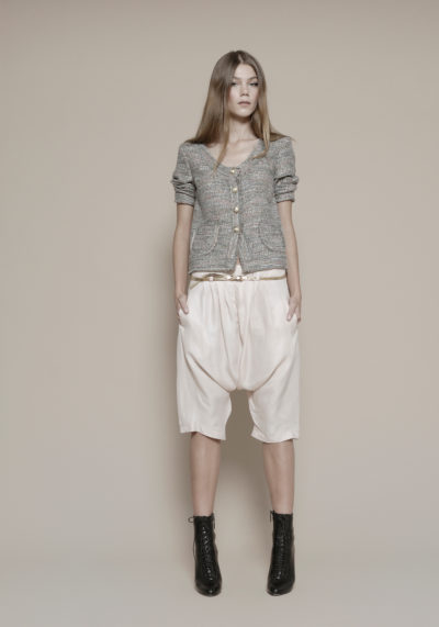 Une mannequin aux cheveux longs se tient debout sur un fond beige uni, photographiée par le photographe Thomas Duclert. Elle porte un cardigan gris à boutons, un short bouffant rose clair, une ceinture dorée et des bottines noires à lacets. Les mains dans les poches, elle regarde vers l'avant, l'air serein.