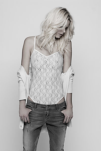 Une mannequin aux cheveux blonds longs et ondulés regarde vers le bas, debout sur un fond clair. Elle porte un caraco en dentelle blanche, un cardigan tombant sur ses épaules et un jean usé. Capture par le célèbre photographe Thomas Duclert, sa tenue respire l'élégance sans effort et le chic moderne.