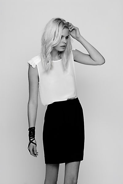 Une mannequin aux cheveux longs et ondulés se tient debout sur un fond uni. Elle porte un chemisier blanc sans manches et une jupe noire à taille haute, avec un bracelet noir au poignet gauche. Sa main droite touche ses cheveux tandis qu'elle regarde légèrement vers le bas. Prise par Thomas Duclert à Paris, l'image respire la mode intemporelle en noir et blanc.