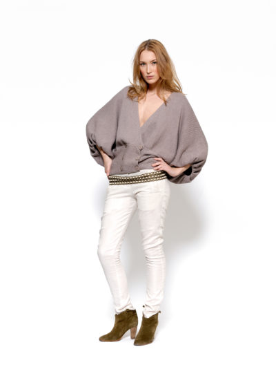 Une mannequin aux cheveux longs et ondulés se tient debout sur un fond blanc. Elle porte un pull gris ample à col en V sur une ceinture cloutée et un pantalon blanc, associé à des bottines marron. Ses mains sont sur ses hanches et elle regarde l'objectif, comme si elle posait pour un photographe à Paris sous la direction de Thomas Duclert.