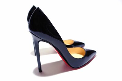 Une paire de chaussures Louboutin à talons hauts en cuir verni noir avec bouts pointus et semelles rouges distinctives. Les chaussures ont une finition élégante et brillante et sont positionnées avec une chaussure légèrement devant l'autre sur un fond blanc, capturant l'essence de la mode parisienne comme si elles avaient été photographiées par un photographe de renom.