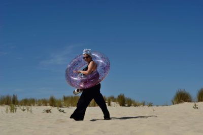 Une personne portant un pantalon noir et un foulard argenté métallique marche sur une plage de sable, tenant une bouée gonflable violette brillante et transparente avec des paillettes. Photographiée par Thomas Duclert de Paris, la scène présente un ciel bleu clair et des herbes vertes poussant dans le sable en arrière-plan.