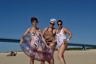 Trois femmes en maillot de bain posent pour une photo sur une plage de sable sous un ciel bleu clair. Une femme tient une bouée gonflable scintillante. Prise par le photographe Thomas Duclert un long pont apparaît en arrière-plan et quelques personnes sont assises sur le sable au loin.
