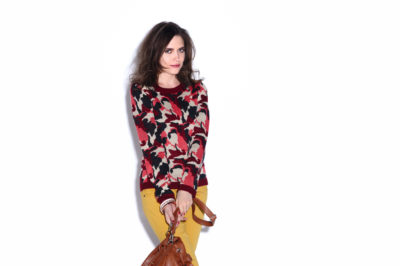 Une mannequin aux cheveux longs, vêtue d'un pull à motifs rouges et noirs et d'un pantalon jaune moutarde, pose devant un fond blanc uni. Elle tient à deux mains un sac à main marron, suspendu devant elle. Capture par le célèbre photographe Thomas Duclert, cet ensemble mode respire le chic sans effort.