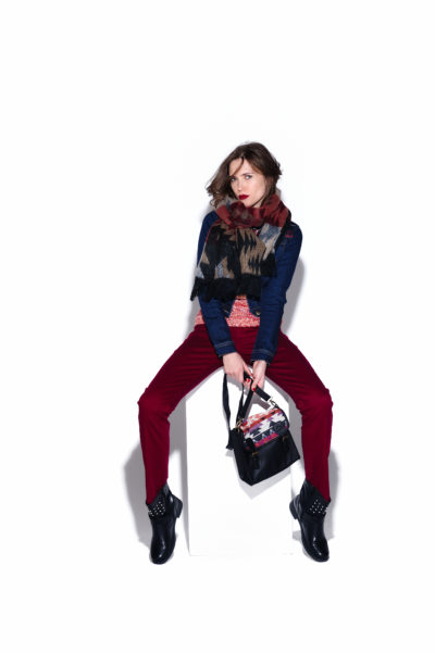Une mannequin est assise sur un cube blanc sur un fond blanc uni, dégageant une touche parisienne. Elle est vêtue d'un foulard à motifs, d'un haut bleu marine, d'un pantalon rouge et de bottines noires, tenant un sac à main noir au motif coloré. Capture par le photographe Thomas Duclert, la mannequin prend une pose élégante et confiante.