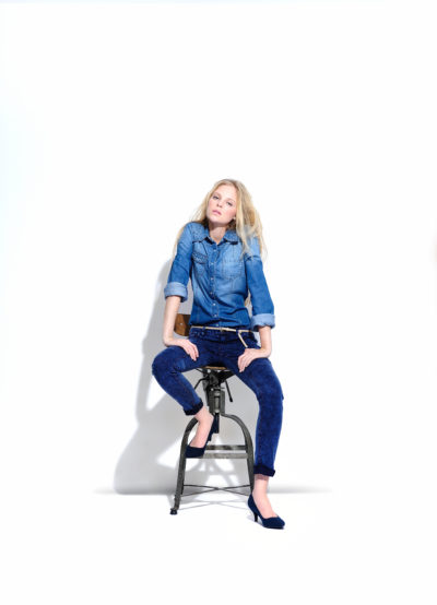 Une mannequin aux longs cheveux blonds est assise sur un tabouret haut sur un fond blanc uni. Elle porte une chemise en jean et un jean bleu foncé, associés à des chaussures bleues. Ses mains reposent sur ses cuisses tandis qu'elle regarde directement l'objectif, incarnant l'esthétique mode de Thomas Duclert dans cette photographie saisissante.
