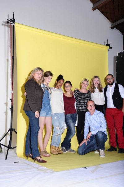 Huit personnes posent pour une photo de groupe sur fond jaune. Thomas Duclert se tient parmi les quatre personnes du milieu, ajoutant une touche de charme parisien. Trois personnes se tiennent à gauche et une personne à genoux à droite. Ils sont habillés de manière décontractée et semi-décontractée, sourient et regardent l'objectif.