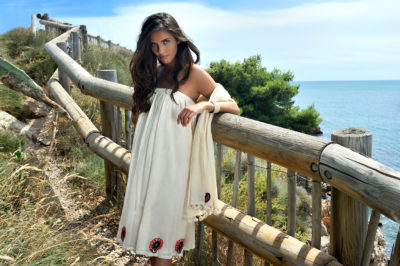 Une mannequin aux longs cheveux noirs se tient debout près d'une barrière en bois près d'un sentier côtier avec la mer visible en arrière-plan. Elle porte une robe blanche à épaules dénudées avec des motifs décoratifs près de l'ourlet. Dans ce décor ensoleillé et pittoresque, on se croirait dans une séance photo de mode tout droit sortie de Paris.