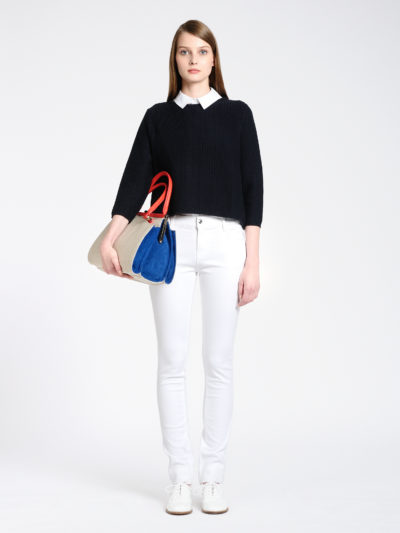 Une mannequin se tient debout sur un fond uni, vêtue d'un pull noir sur une chemise blanche à col et d'un pantalon blanc. Elle porte un sac élégant aux couleurs bleues, beiges et rouges. Ses cheveux raides tombent sur ses épaules et elle porte des chaussures blanches, une parfaite capture de la mode par le photographe Thomas Duclert.