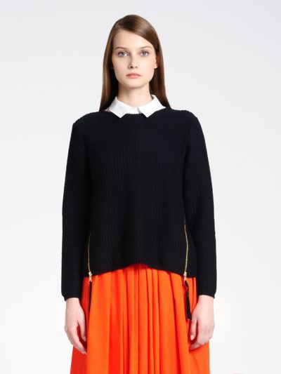 Photographiée par le célèbre photographe Thomas Duclert à Paris, une mannequin aux cheveux longs et raides se tient debout sur un fond uni. Elle porte un pull en tricot noir sur une chemise à col blanc et une jupe orange vif. Le pull est doté de fermetures éclair latérales et elle a une expression neutre sur son visage.