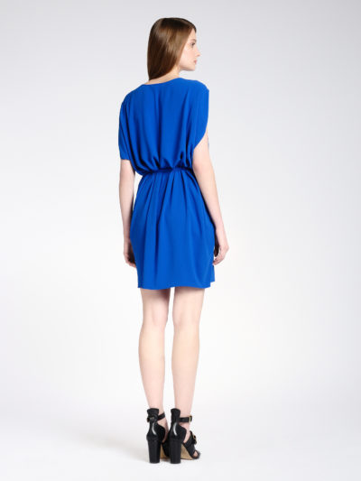 Une mannequin aux longs cheveux bruns se tient debout, le dos tourné, vêtue d'une robe bleue ample jusqu'aux genoux avec une taille froncée. Elle porte également des sandales noires à talons hauts. L'arrière-plan est un décor de studio blanc uni, évoquant l'élégance minimaliste souvent observée dans la photographie de mode parisienne.